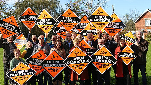 A large group of Liberal Democrats in the sun holding signs that say Liberal Democrats Winning Here